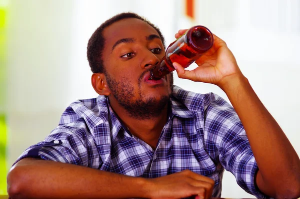 Homem bonito vestindo camisa azul branco sentado ao balcão do bar deitado sobre a mesa bebendo de garrafa de cerveja marrom, bêbado expressão facial deprimida, conceito alcoólico — Fotografia de Stock