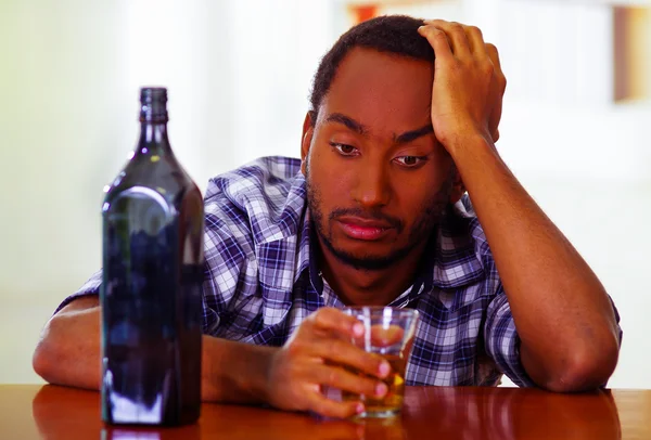 Homme portant une chemise bleue blanche assise près d'un comptoir de bar tenant un verre de whisky à côté d'une bouteille d'alcool, expression faciale déprimée — Photo
