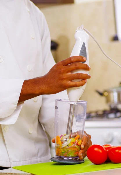 Detailní záběr rukou šéfkuchaře střídat barevné zeleniny do průhledného skla pomocí ruční mixér, profesionální kuchyně, bílé pozadí — Stock fotografie