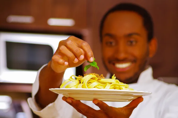 Chef satisfait portant des vêtements blancs plaçant la feuille verte sur le dessus du plat de spaghetti, souriant heureux tenant la plaque dans l'air pour la caméra — Photo