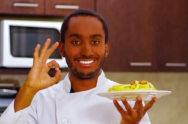 Satisfied chef wearing white clothes making circle with fingers expressing exquisite taste, smiling happily holding plate up in air for camera — Stock Photo, Image