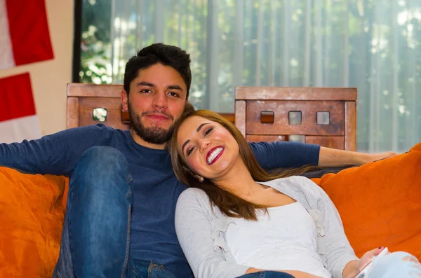 Joven pareja encantadora sentada y abrazándose en un sofá naranja sonriendo a la cámara, ambiente del albergue — Foto de Stock