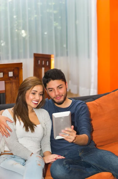 Jovem casal encantador sentado e abraçando assistindo tela tablet no sofá laranja sorrindo para a câmera, ambiente albergue — Fotografia de Stock