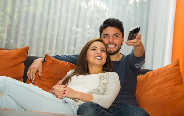 Joven pareja encantadora sentada en un sofá naranja abrazando y viendo la televisión, él apuntando el control remoto hacia la cámara, el entorno del albergue —  Fotos de Stock