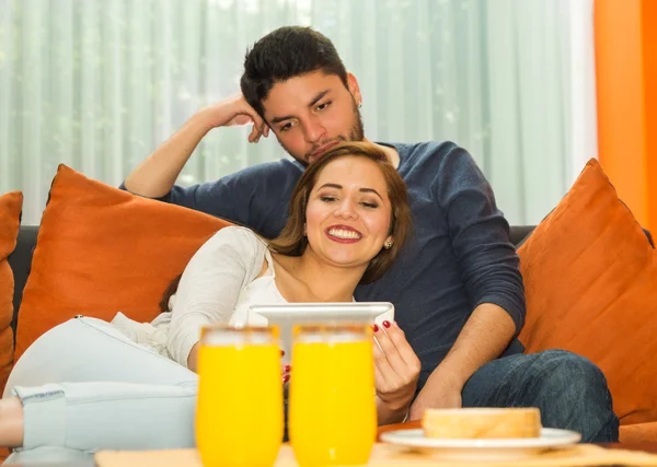 Молодая очаровательная пара сидит и обнимает смотреть планшетный экран в оранжевый диван улыбаясь в камеру, общежитие среде — стоковое фото