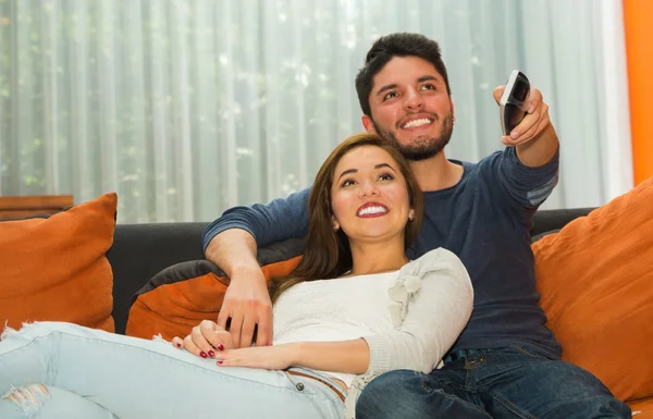 Young charming couple seated in orange sofa embracing and watching television, him pointing remote control towards camera, hostel environment — Stock Photo, Image