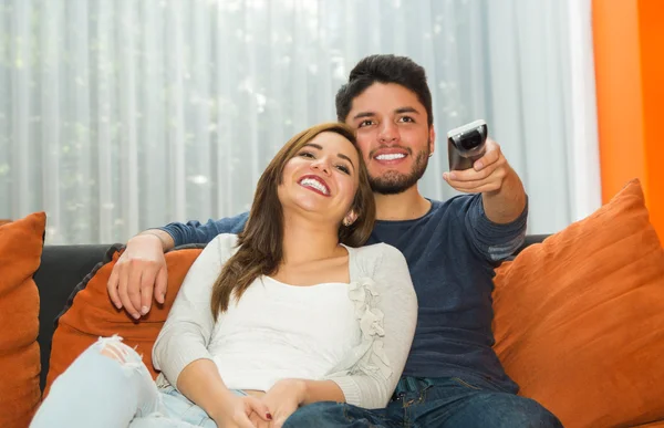 Jovem casal encantador sentado no sofá laranja abraçando e assistindo televisão, ele apontando controle remoto para câmera, ambiente albergue — Fotografia de Stock