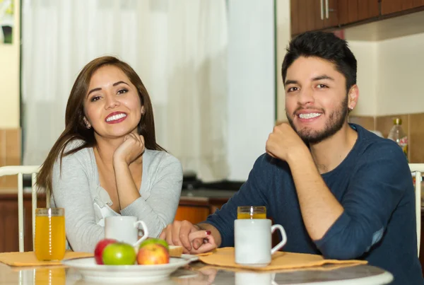 Jeune couple charmant assis près de la table de petit déjeuner souriant à la caméra, fruits, jus et café placés devant, environnement de l'auberge — Photo
