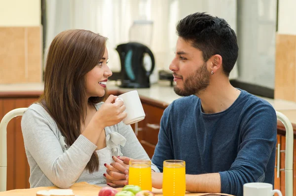 Jonge charmante paar gezeten door ontbijttafel glimlachen naar de camera, fruit, SAP en koffie geplaatst vooraan, hostel milieu — Stockfoto