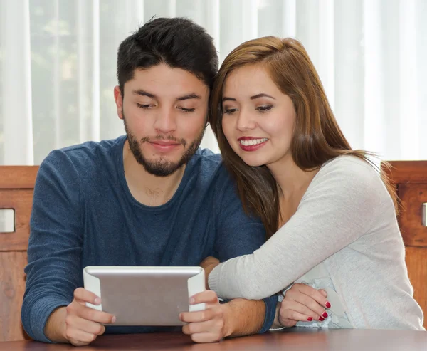 Junges charmantes Paar am Tisch sitzend und Tablet-Bildschirm beobachtend, während es sich umarmt, glücklich und lächelnd, Herbergskonzept — Stockfoto