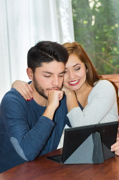 Joven pareja encantadora sentada junto a la mesa viendo la pantalla de la tableta mientras abraza, bot feliz y sonriente, concepto de albergue —  Fotos de Stock