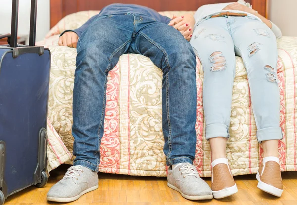 Closeup pernas casal vestindo jeans, sentado na borda da cama, mala azul de pé ao lado, conceito albergue — Fotografia de Stock