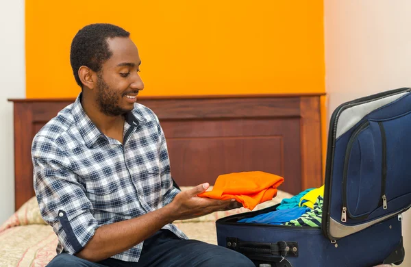 Beau jeune homme assis sur le lit valise d'emballage et souriant, concept invité auberge — Photo