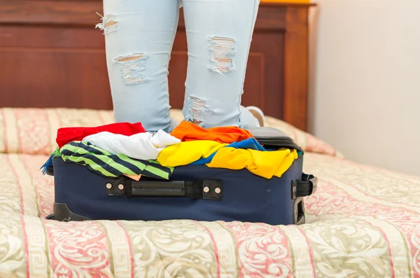 Öppen resväska med kläder inuti liggande på sängen, kvinnans ben i bakgrunden, hostel pensionat koncept — Stockfoto