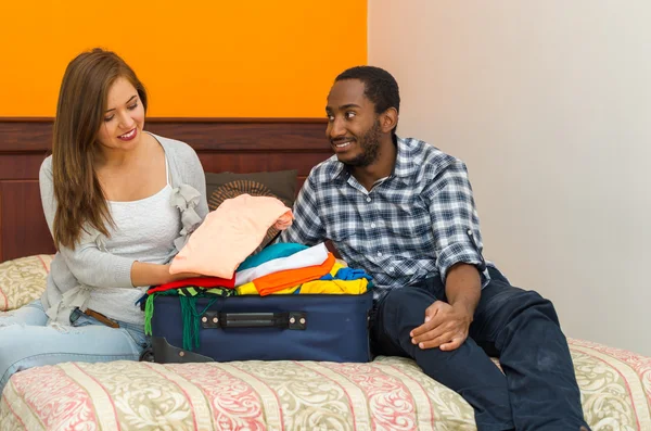 Encantadora pareja joven sentada en la cama interactuando felizmente mientras empaca la maleta con la ropa, concepto de huésped del albergue — Foto de Stock