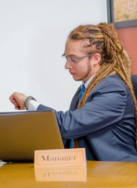 Handsome man with dreads and business suit sitting by desk looking at wrist watch, young manager concept — Stock Photo, Image