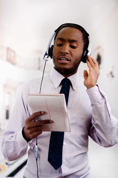 Knappe man dragen hoofdtelefoon met microfoon, wit gestreept shirt en stropdas, poseren houder notebook in de hand, lachende interactie — Stockfoto