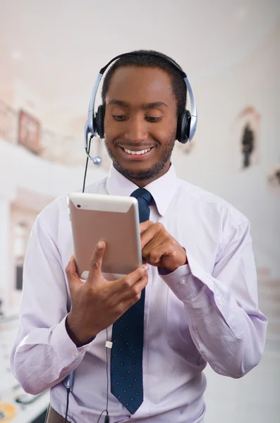 Beau homme portant des écouteurs avec microphone, chemise à rayures blanches et cravate, posant tenant la tablette à la main, souriant interagissant — Photo