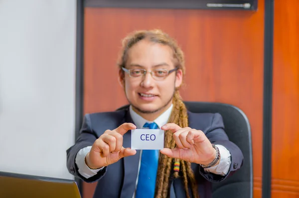 Handsome man with dreads, glasses and business suit sitting by desk holding up a paper which has the word executive written on it in his pocket, young manager concept — Stock Photo, Image