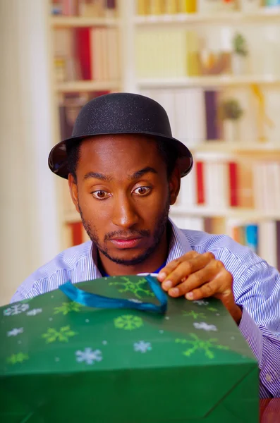 Charmant homme portant chemise bleue et chapeau assis à la table d'ouverture cadeau d'anniversaire, l'air légèrement excité, célébrer concept seul — Photo