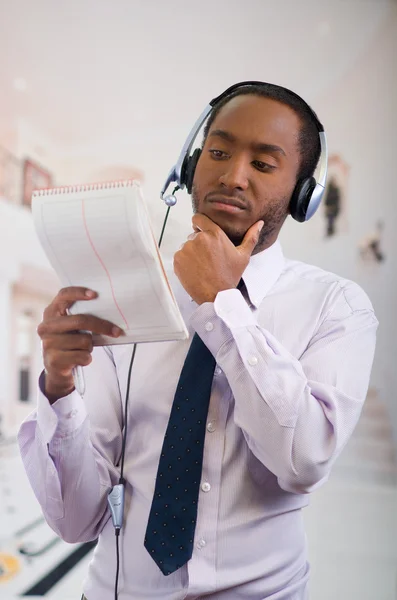 Hombre guapo usando auriculares con micrófono, camisa blanca a rayas y corbata, posando sosteniendo el cuaderno en la mano, sonriendo interactuando —  Fotos de Stock