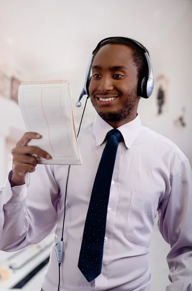 Beau homme portant des écouteurs avec microphone, chemise à rayures blanches et cravate, posant tenant un cahier à la main, souriant interagissant — Photo