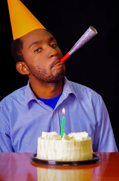 Gut aussehender Mann in blauem Hemd und Hut am Tisch sitzend mit Kuchen davor, Kerze brennend, Partyhorn blies vor laufender Kamera, allein zelebrierend Konzept — Stockfoto