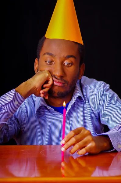 Homme à l'air ennuyé portant chemise bleue et chapeau assis à table tenant une seule bougie brûlante, triste expression face à la caméra, célébrant concept seul — Photo