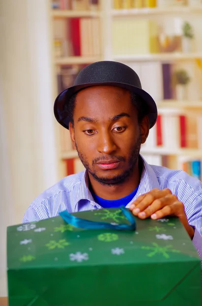 Charmant homme portant chemise bleue et chapeau assis à la table d'ouverture cadeau d'anniversaire, l'air légèrement excité, célébrer concept seul — Photo