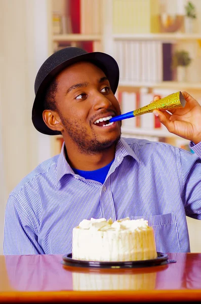 Очаровательный мужчина в голубой рубашке и шляпе сидит за столом с праздничным тортом перед носом, трубит в рог, празднуя в одиночестве улыбаясь — стоковое фото