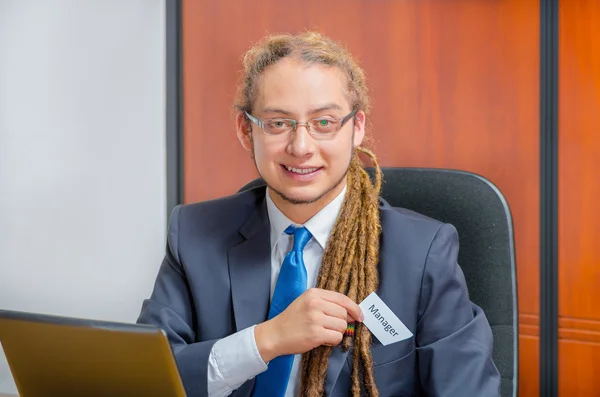 Handsome man with dreads, glasses and business suit sitting by desk placing a paper which has the word ceo written on it in his pocket, young executive concept — Stock Photo, Image