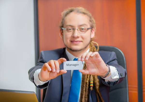 Handsome man with dreads, glasses and business suit sitting by desk holding up a paper which has the word manager written on it in his pocket, young executive concept — Stock Photo, Image