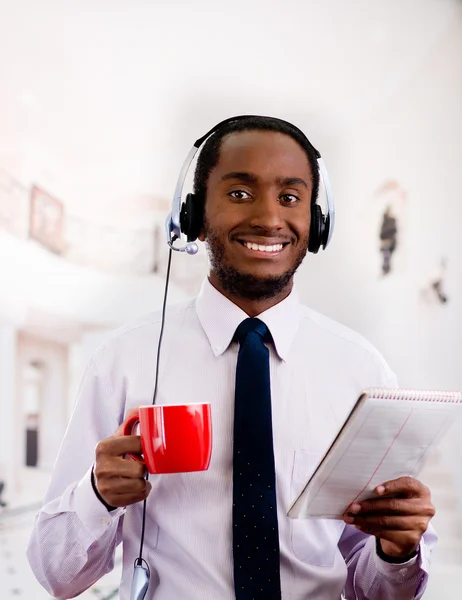 Beau homme portant un casque avec microphone, chemise à rayures blanches et cravate, tenant une tasse de café, souriant à la caméra, concept d'entreprise — Photo