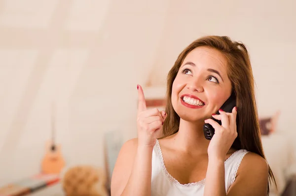 Jeune femme attrayante portant un haut blanc parlant au téléphone tout en souriant, caméra face — Photo