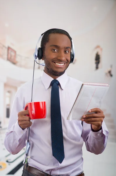 Beau homme portant un casque avec microphone, chemise à rayures blanches et cravate, tenant une tasse de café, souriant à la caméra, concept d'entreprise — Photo