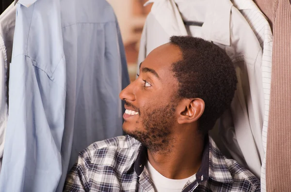 Headshot jeune homme souriant à la caméra, assis avec des chemises suspendues autour de sa tête, concept de mode — Photo