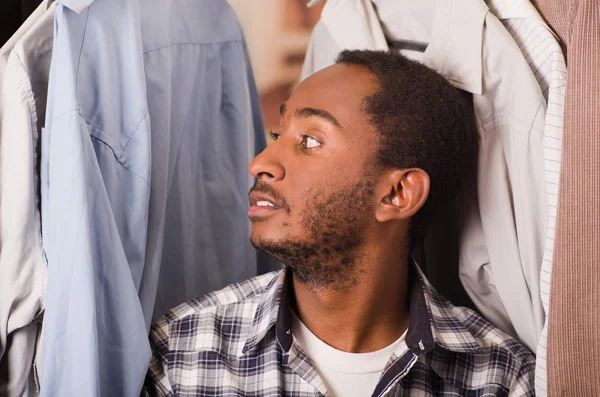 Cabeza joven sonriendo a la cámara, sentado con camisas colgando alrededor de su cabeza, concepto de moda — Foto de Stock