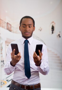 Stresli yüz ifadesi ile iki cep telefonu tutan gömlek ve kravat giyen yakışıklı adam