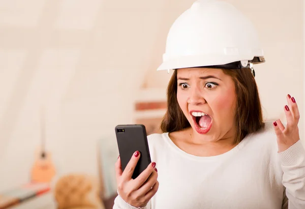 Jeune femme portant un casque de construction regardant le téléphone portable avec expression faciale stressée — Photo