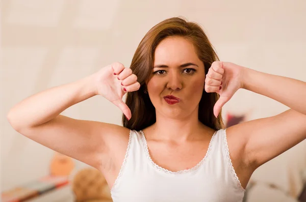 Mujer joven con la parte superior blanca y auriculares frente a la cámara mientras interactúa frustración mostrando dos pulgares hacia abajo — Foto de Stock