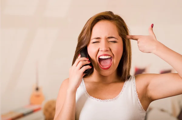 Jonge vrouw draagt witte top geconfronteerd camera terwijl interactie frustratie praten op de telefoon, het maken van pistool met vingers gericht op haar eigen hoofd — Stockfoto