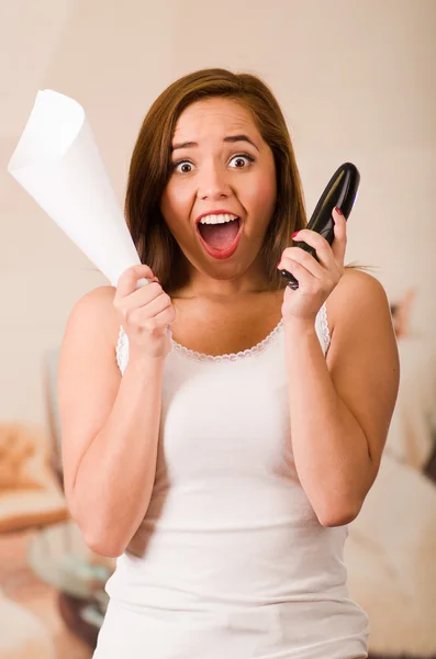 Young woman wearing white top facing camera while screaming in frustration, holding paper and mobile phone, stressed concept — Stock Photo, Image