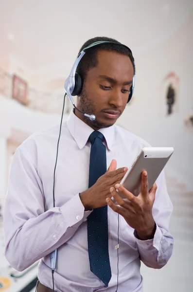 Beau homme portant des écouteurs avec microphone, chemise à rayures blanches et cravate, posant tenant la tablette à la main, souriant interagissant — Photo