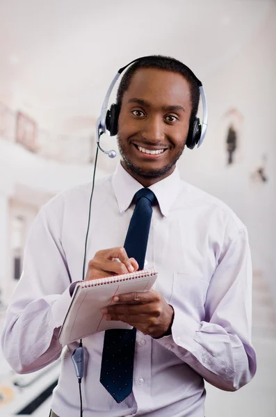 Beau homme portant des écouteurs avec microphone, chemise à rayures blanches et cravate, posant tenant un cahier à la main, souriant interagissant — Photo