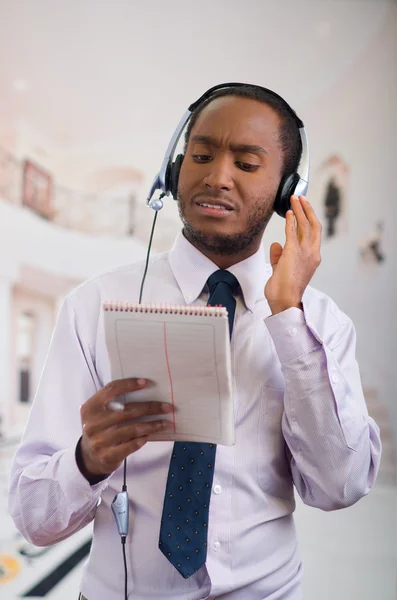 Beau homme portant des écouteurs avec microphone, chemise à rayures blanches et cravate, posant tenant un cahier à la main, souriant interagissant — Photo