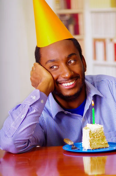 Очаровательный мужчина в голубой рубашке и шляпе сидит за столом с кусочком торта впереди, выглядит счастливым, празднуя одинокую концепцию — стоковое фото