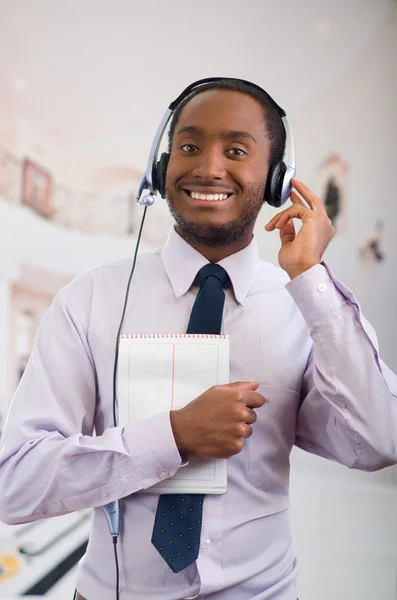 Beau homme portant des écouteurs avec microphone, chemise à rayures blanches et cravate, posant tenant un cahier à la main, souriant interagissant — Photo