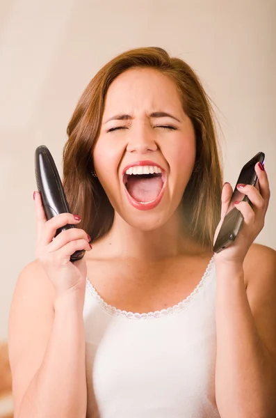 Jonge vrouw draagt witte top geconfronteerd camera terwijl schreeuwen in frustratie, met twee telefoons, gestrest concept — Stockfoto