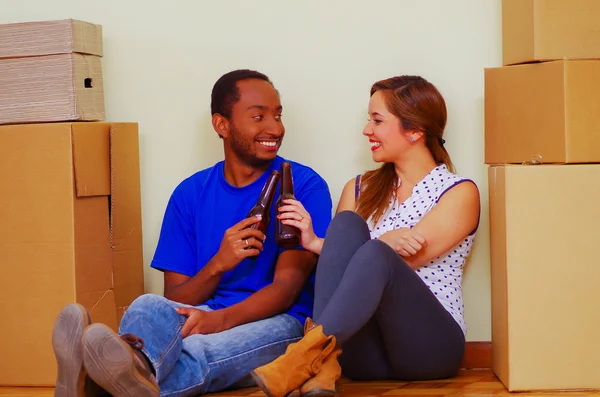 Encantadora pareja interracial sonriendo a la cámara, amigable interactuando mientras está sentado entre cajas de cartón, moviéndose en concepto — Foto de Stock