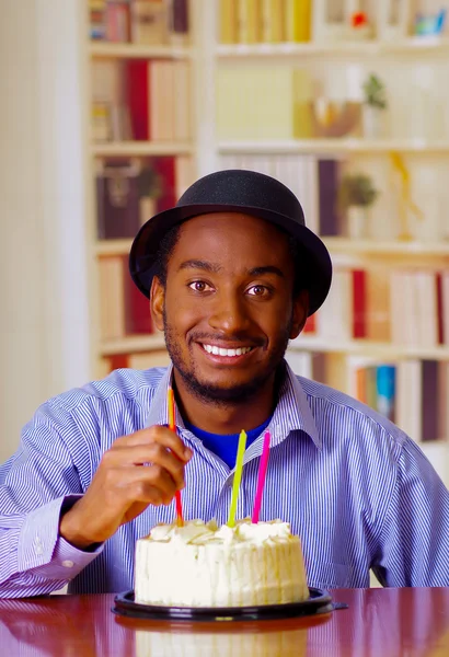 Charmant homme portant une chemise bleue et un chapeau assis à table avec un gâteau d'anniversaire devant, touchant une bougie célébrant seul en souriant — Photo